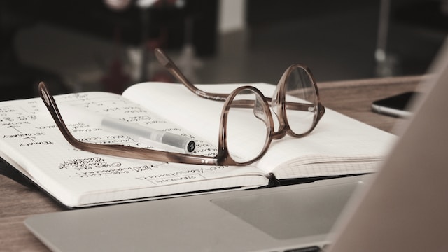 Photo of a pair of glasses ontop of a notebook next to a laptop by Dan Dimmock on Unsplash