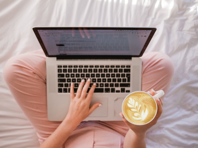 Photo of a person sitting on a bed using a laptop and holding a coffee by Sincerely Media on Unsplash